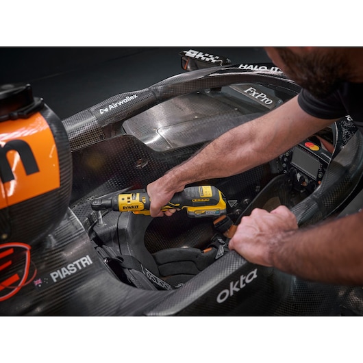 A DEWALT DCF514N cordless ratchet tool is being used inside a race car cockpit, with a person's hands operating the tool on a metal component. The cockpit and several sponsor logos are visible.