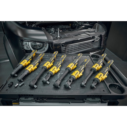 Several DEWALT cordless ratchets are displayed on a black workbench in front of a car with its hood open. The ratchets are yellow and black, featuring different head types, and are arranged in a row. Some socket attachments are also visible on the workbench.