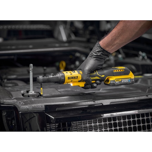 A DEWALT DCF520N cordless ratchet tool being used by a person wearing black gloves to tighten a bolt on a car engine. The tool features an 18V battery and Power Stack technology.