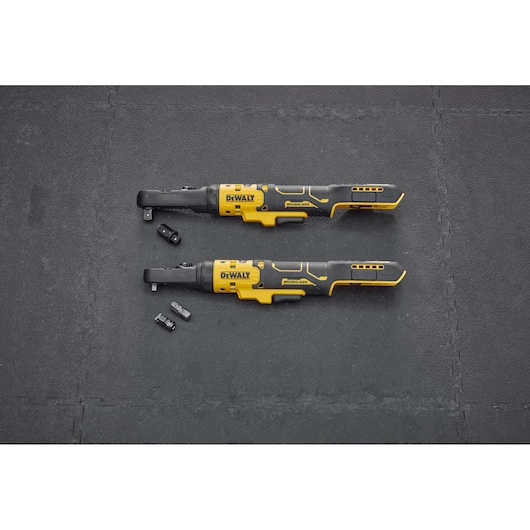 Two DEWALT cordless ratchet wrenches with interchangeable attachments are placed on a black textured surface. The tools are yellow and black, and several small accessory parts are positioned nearby.