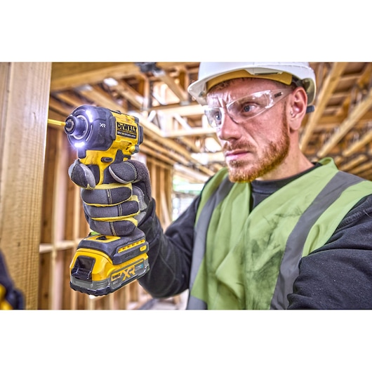 A person wearing a high-visibility vest and work gloves uses a DEWALT cordless impact driver with LED light on a construction site. The face of the person is blurred for privacy.