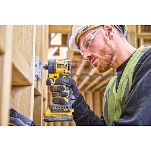 A person wearing gloves, a safety vest, and a hard hat is using a DEWALT DCF870E2T cordless impact driver to fasten a metal bracket onto a wooden construction frame.