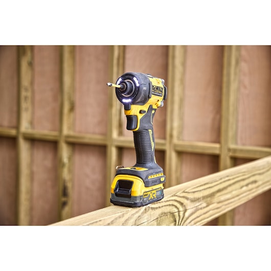 A DEWALT cordless impact driver, model DCF870E2T, sitting upright on a wooden beam at a construction site. The tool has a yellow and black design and appears to be turned on, with LED lights illuminated around the drill bit.
