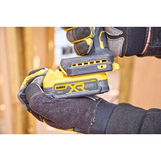 Close-up of gloved hands holding a DEWALT 18V XR Power Stack cordless tool, highlighting the battery pack and tool grip.