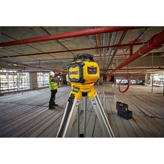A DEWALT DCLE05211R laser level mounted on a tripod in an unfinished building construction site, with a worker in a safety vest and helmet measuring or aligning pipes in the background.