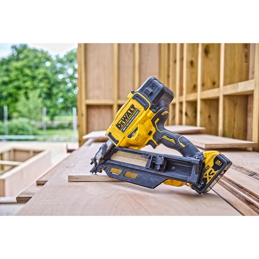 8V XR Brushless framing nailer stands on top of a wooden surface in an open construction site. Trees are visible in the background