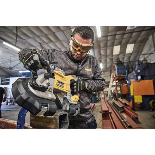 A person wearing protective gloves and clothing is using a DEWALT DCS374N portable band saw to cut a piece of metal in an industrial workshop. The person's face is blurred for privacy.