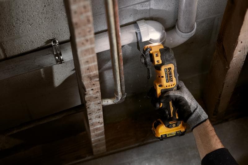 Person uses a DEWALT cordless oscillating tool to cut a pipe against a concrete wall in a construction setting.