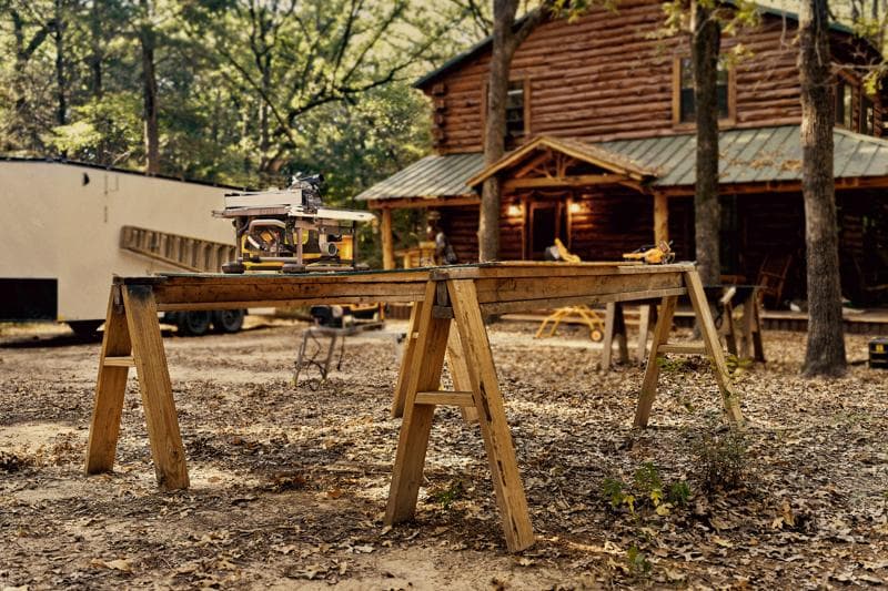 A DEWALT table saw set up on a wooden workbench made from two sawhorses outdoors, with a log cabin and trees in the background.