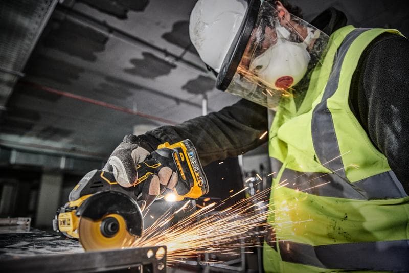 A person wearing safety gear, including a helmet, face shield, mask, gloves, and a high-visibility vest, is using a DEWALT DCS438N cordless power tool to cut metal, creating sparks.