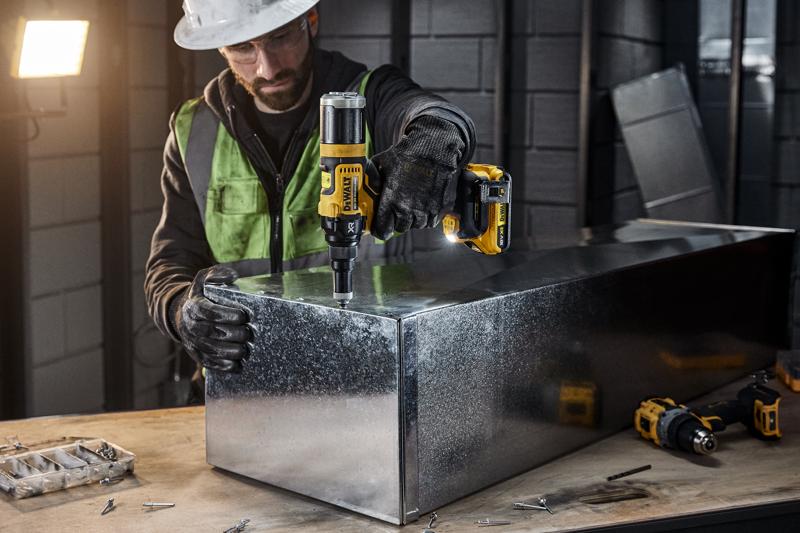 A person wearing safety gear uses a DEWALT DCF403NT cordless power tool to work on a metal box in an industrial workshop. Various screws and tools are visible on the workbench.