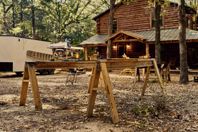 A DEWALT table saw set up on a wooden workbench made from two sawhorses outdoors, with a log cabin and trees in the background.