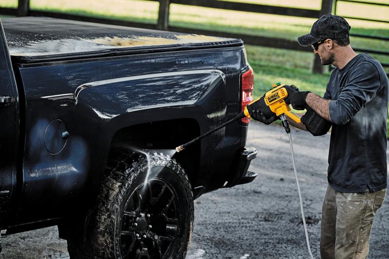 A person is using a DEWALT DCPW550B cordless power washer to clean the wheel and side of a black pickup truck outdoors.
